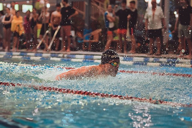 A swimmer performs the breaststroke during an indoor swimming competition, with water splashing around and spectators watching in the background.